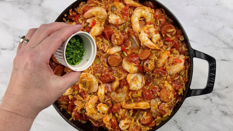 hand hovering chopped chives over a pot of vegetables, pasta, sausage, and shrimp