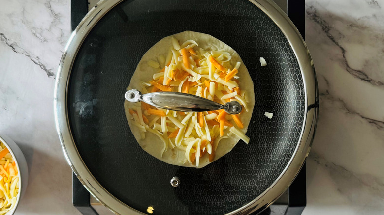 flour tortilla topped with shredded cheese in a lidded frying pan