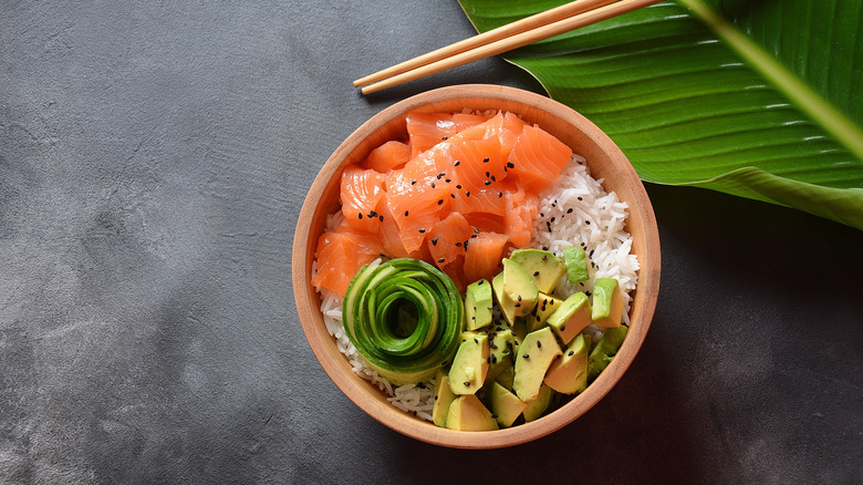 poke bowl with salmon, avocado and black sesame seeds; leaf and bamboo on table with poke bowl