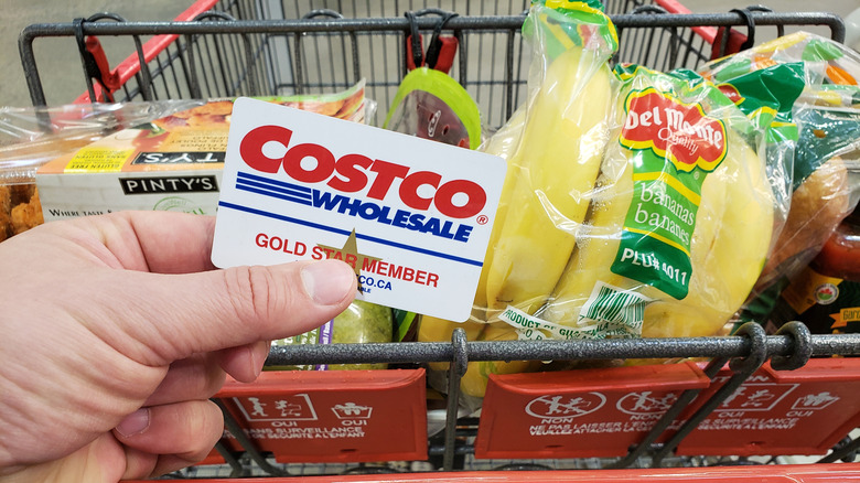 Person holding a Costco membership card over a shopping cart full of groceries.