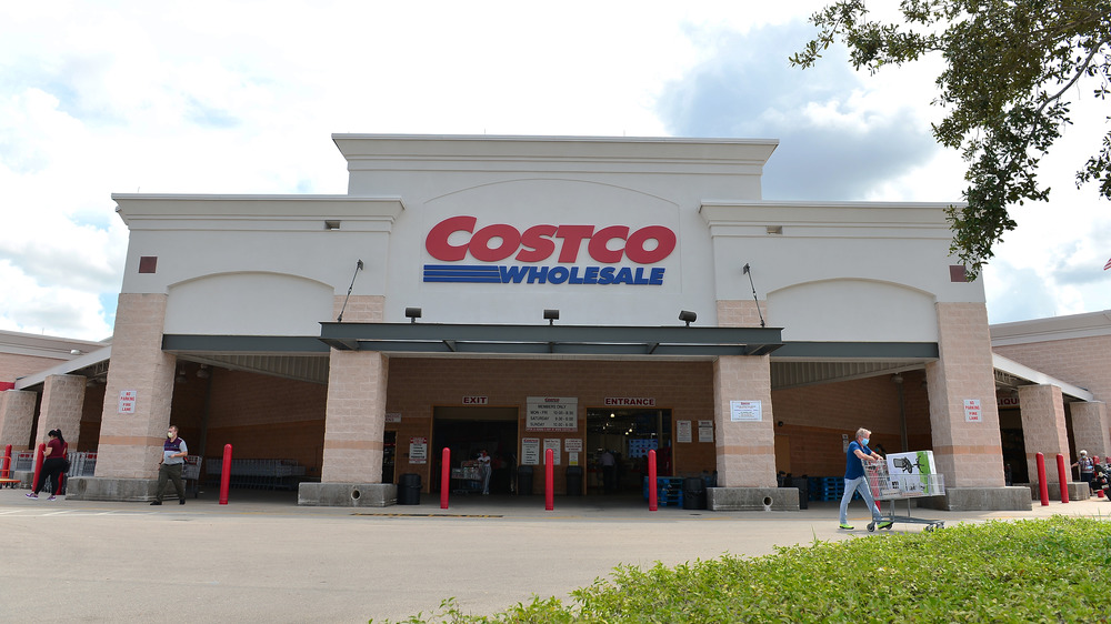 A giant Costco rears over a diminutive masked shopper as they cart away their boxed chair, the gift of the changeable savings brand.