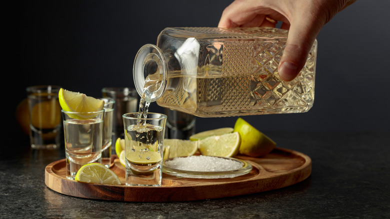 A hand pouring tequila out of a bottle with shot glasses, lime wedges, and salt on wood platter.