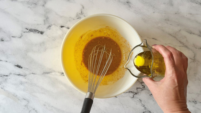Hand pouring olive oil into bowl with dressing and whisk