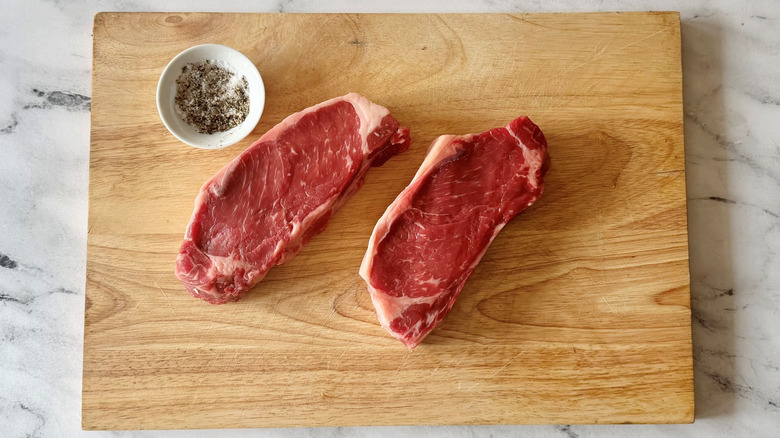 Raw steaks on wooden board with small bowl of salt and pepper