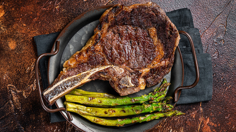 A cooked bone-in ribeye sits on a pan with a side of roasted asparagus.