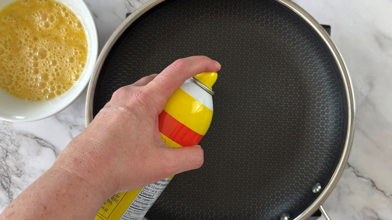 hand squirting cooking spray on frying pan next to bowl of beaten eggs