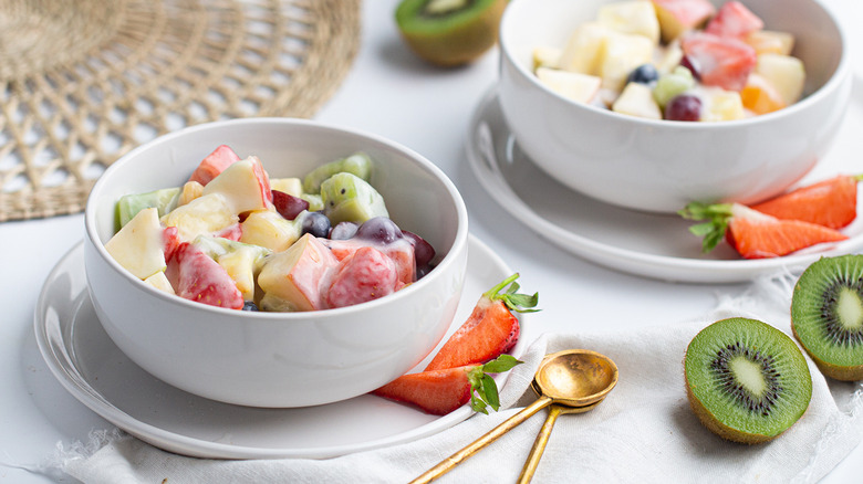 Two white bowls of fruit salad on two small white plates next to strawberries, kiwis, and gold spoons