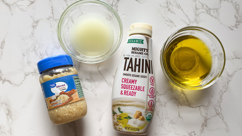 lemon juice in a glass bowl, olive oil in a glass bowl, jar of minced garlic, and a bottle of tahini on a counter