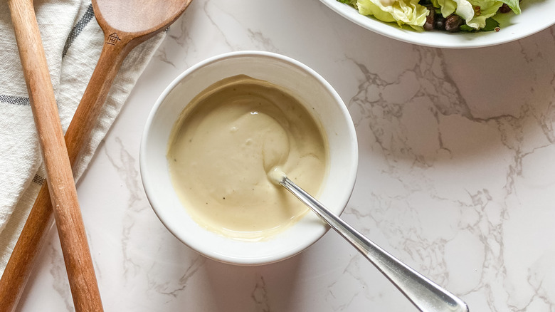 white bowl of creamy lemon tahini dressing near a bowl of greens and wooden spoons on a counter