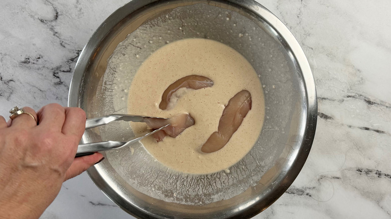 hand holding tongs above a metal bowl filled with raw chicken strips floating in batter