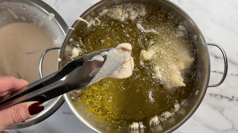 hand holding tongs above a metal pot of battered chicken strips floating in oil