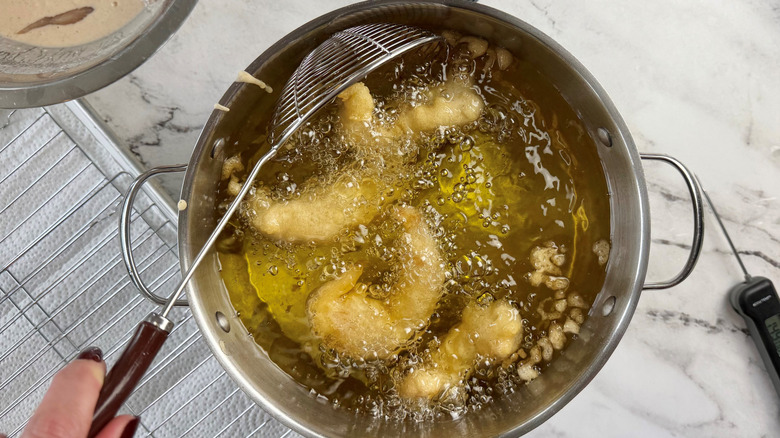 hand holding a metal strainer in a metal pot of battered chicken strips floating in oil