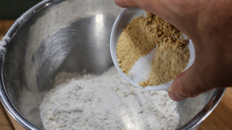hand holding small dish of spices above mixing bowl of flour