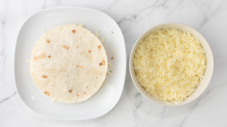 an assembled quesadilla on a white plate next to bowl of cheese