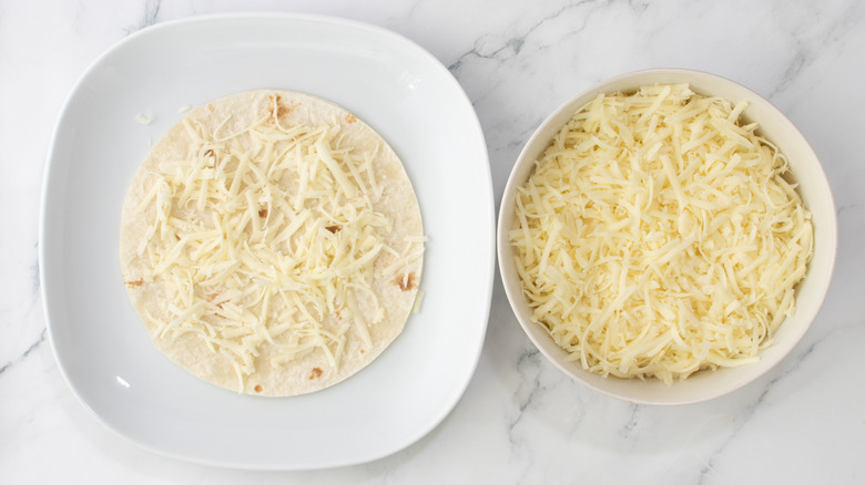 flour tortilla topped with shredded cheese on a white plate next to bowl of cheese
