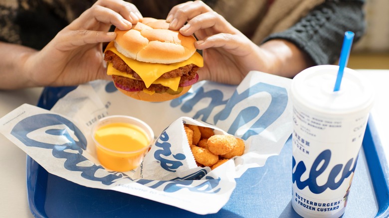Person holding a Culver's burger above fried cheese curds and a drink cup on a tray
