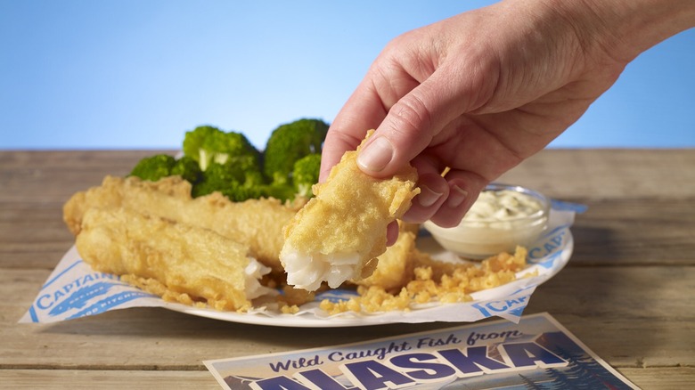 Person holding a breaded fish piece over a plate of food.