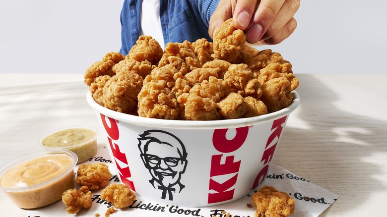 Person taking a piece of popcorn chicken from KFC-branded bucket, with sauces on the side.
