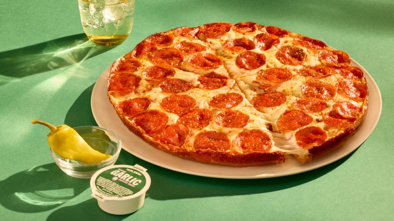 Pan-style Papa Johns pizza on plate next to beverage, garlic dipping sauce, and pepperoncini.