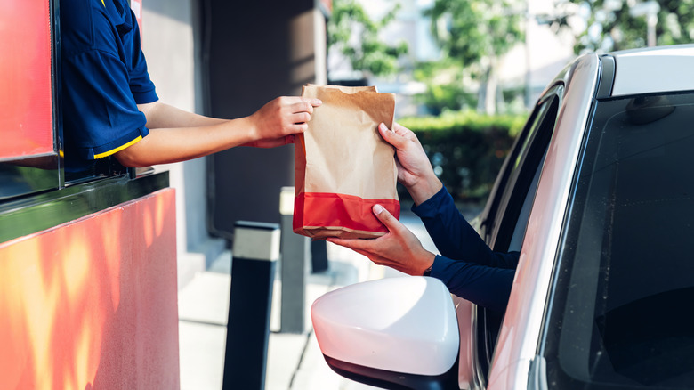 Customer in a white car receives fast food in a brown and red paper sack through a drive thru window.