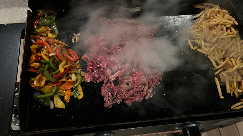 Sizzling piles of veggies, meat, and french fries on the black surface of a Blackstone grill