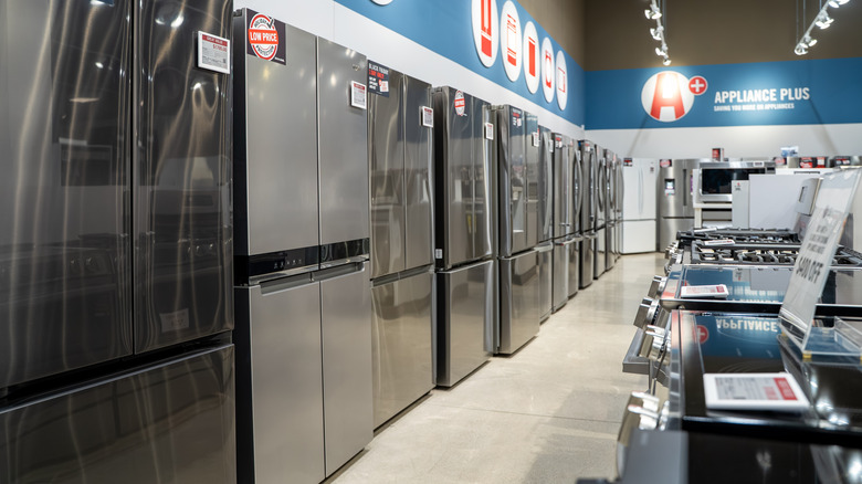 A row of stainless steel refrigerators lining a wall in an appliance store with stoves on the other side.