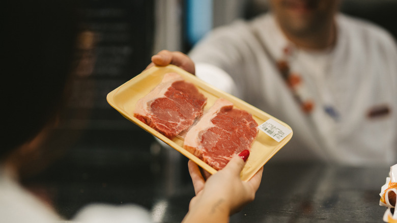 A butcher behind the counter hands over two steaks on a yellow tray and wrapped in clear plastic to a female customer