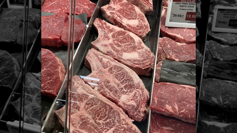 Bone-in prime steaks displayed in a row inside a glass display case, beside other cuts of meat