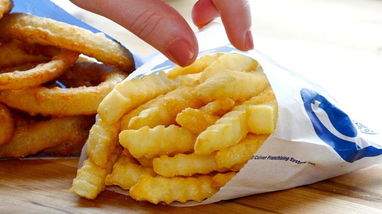 Fingers with painted nails grabbing french fries from Culver's paper bag on wooden tabletop.