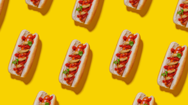Multiple hot dogs topped with vegetables and ketchup in buns arranged over a yellow background