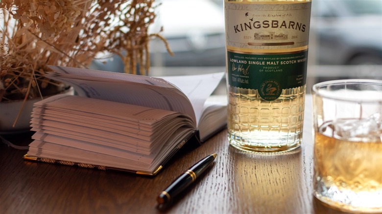 A bottle of Kingsbarns Lowland Scotch whisky on a table, next to a glass, a book, and a pen.
