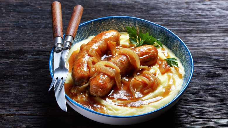 Dish of bangers and mash with onion gravy and a fork and knife on the side of a blue ceramic bowl