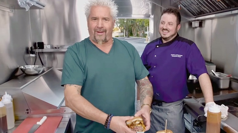 Guy Fieri holding a large hamburger in a food truck with restaurant owner looking on