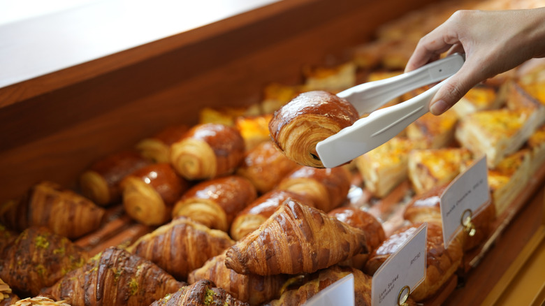 Hand using tongs to pluck chocolate croissant from bakery case.