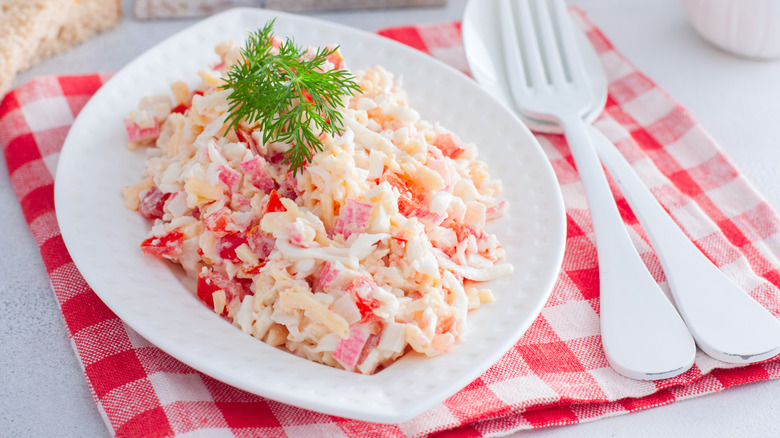 Imitation crab salad in white bowl topped with green herbs on a checkered red napkin with utensils