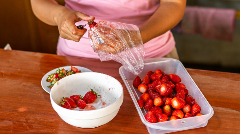 A person cuts the tops off of strawberries, with a bowl below.