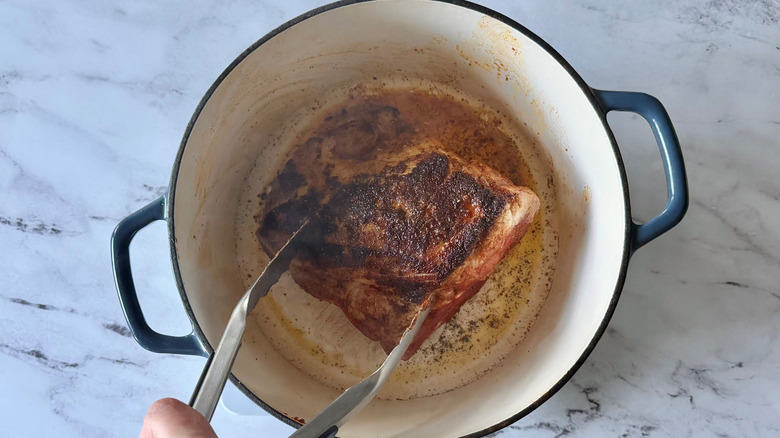 Tongs flipping seared pork in Dutch oven