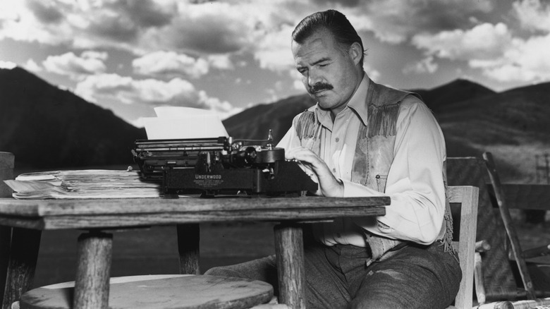 Black-and-white photo of Ernest Hemingway using typewriter at outdoor table.