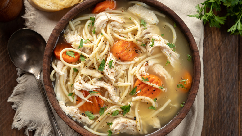 Wooden bowl of chicken noodle soup on table with metal spoon.