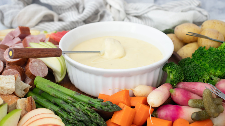 Cheese fondue in bowl surrounded by various vegetable, meat, and fruit dippers