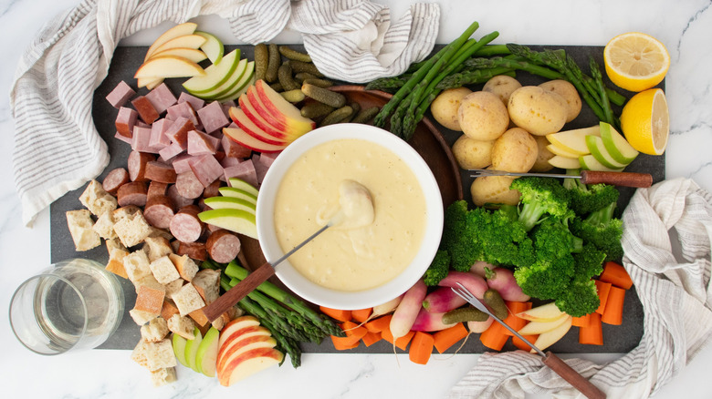 Cheese fondue in bowl surrounded by various vegetable, meat, and fruit toppings