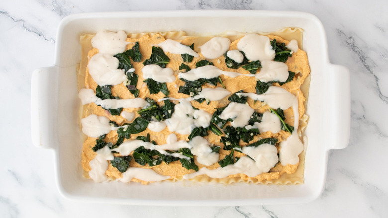 lasagna noodles topped with pumpkin ricotta, sauteed kale, and Alfredo sauce in casserole dish