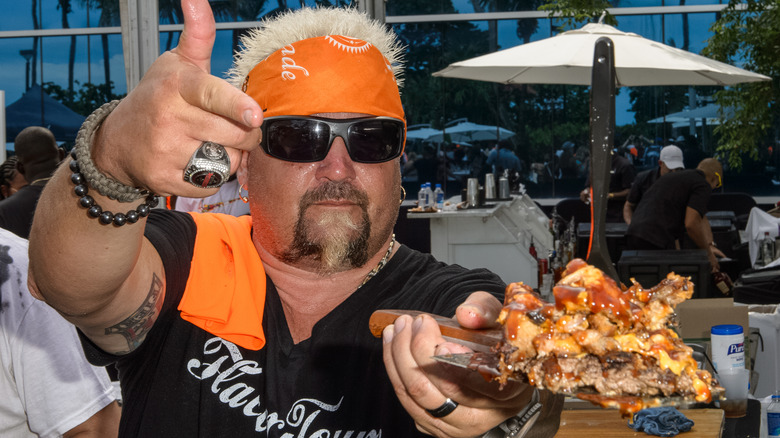 Guy Fieri in orange bandanna, holding up a platter of BBQ