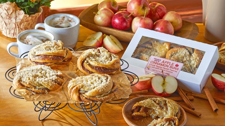 Apple cinnamon buns on metal display next to a box of them, cocoa, fresh apples, and cinnamon sticks