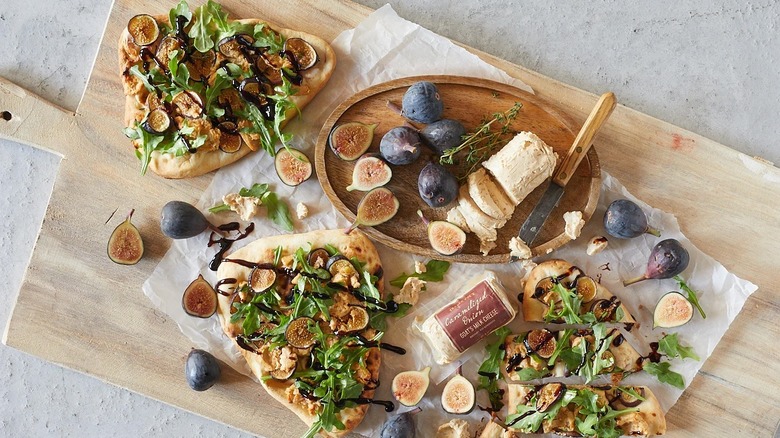 Figs and goat cheese both in and out of packaging, surrounded by fig and arugula flatbread on wood