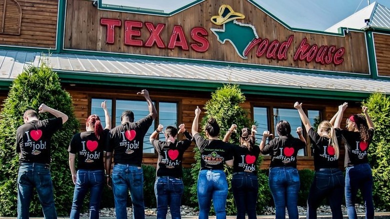Nine Texas Roadhouse employees standing in front of a Texas Roadhouse and pointing to the back of their shirts