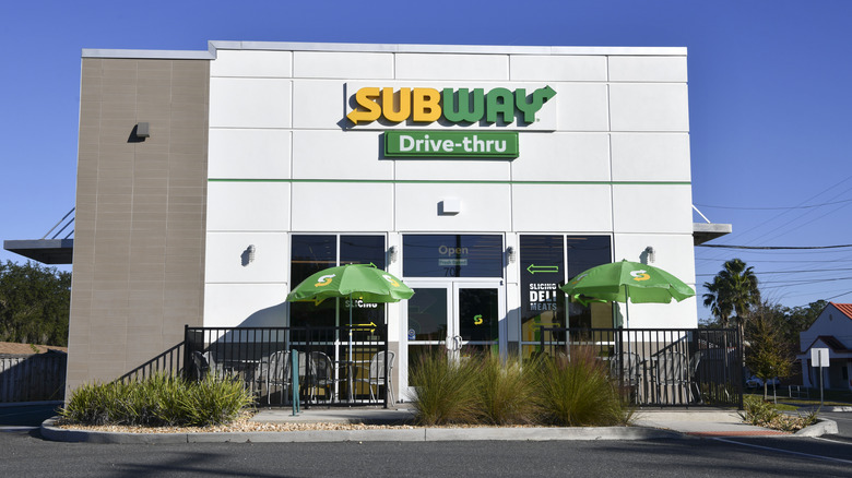 Exterior of a Subway location with a drive-thru, with outdoor seating with green umbrellas.