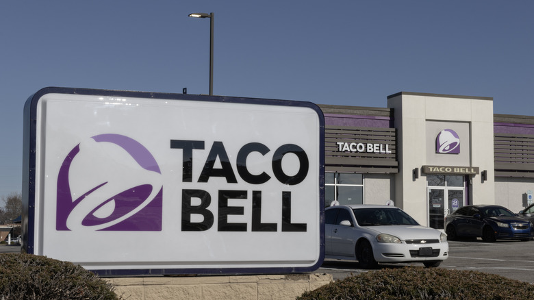 Taco Bell restaurant, with sign in foreground, and one black and one white car parked in lot.