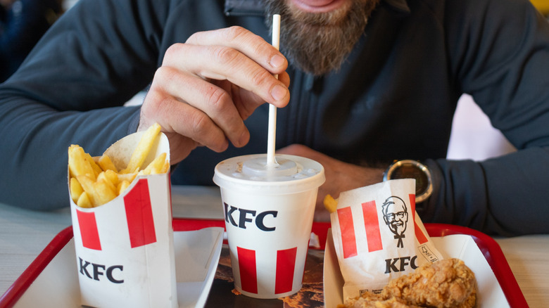 Right hand of bearded man holding straw of KFC drink in the middle of tray of fries, and fried chicken.