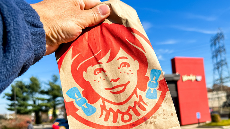 Hand holding Wendy's bag with small grease stains in foreground, with freestanding Wendy's in background.